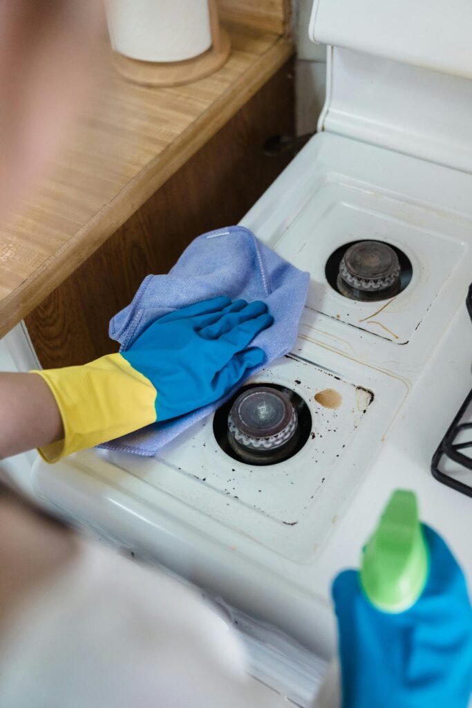 A person wearing rubber gloves cleaning a dirty gas stove with a cloth and spray bottle.