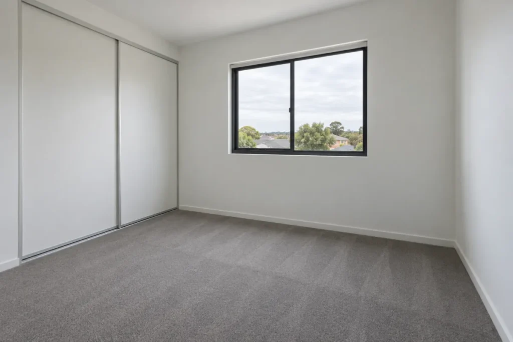 Empty carpeted bedroom with built-in sliding wardrobe and a large window – end of lease cleaning